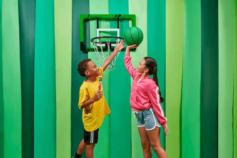 Two children playing basketball, putting a green ball in the hoop with a green backdrop.
