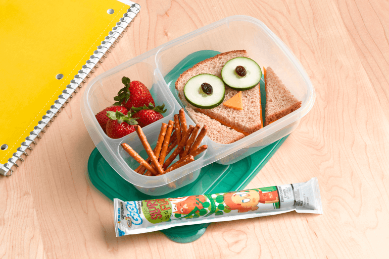 An open lunch box with a sandwich cut into the shape of an owl, strawberries, pretzels and a Go-GURT tube, sitting on a wooden table beside a yellow notepad.